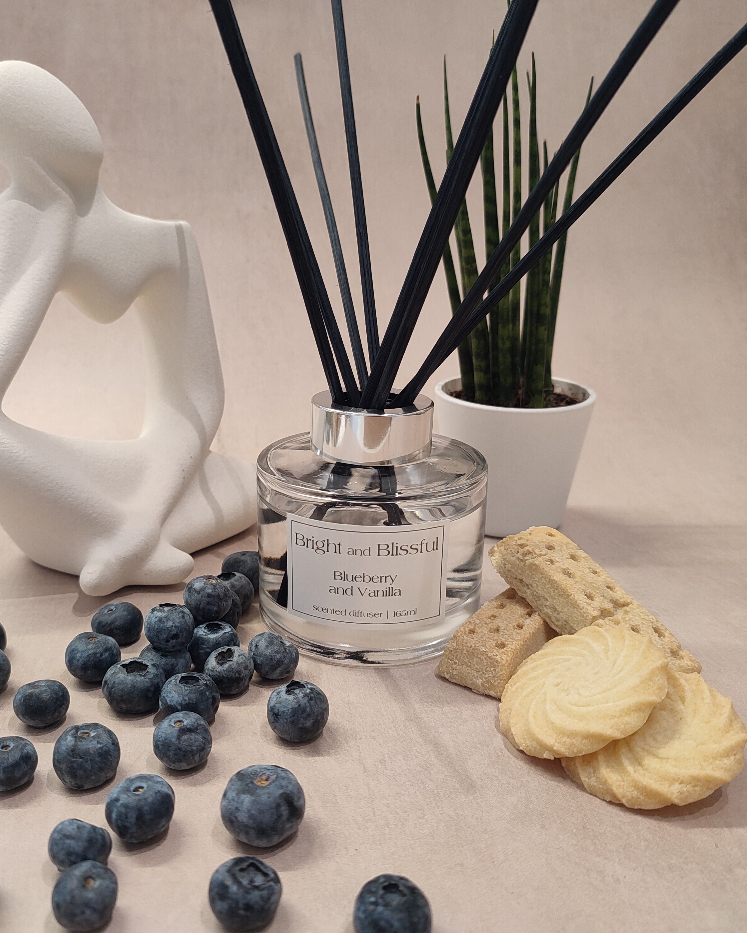 Reed diffuser with blueberries, cookies, and a decorative figure on a neutral background