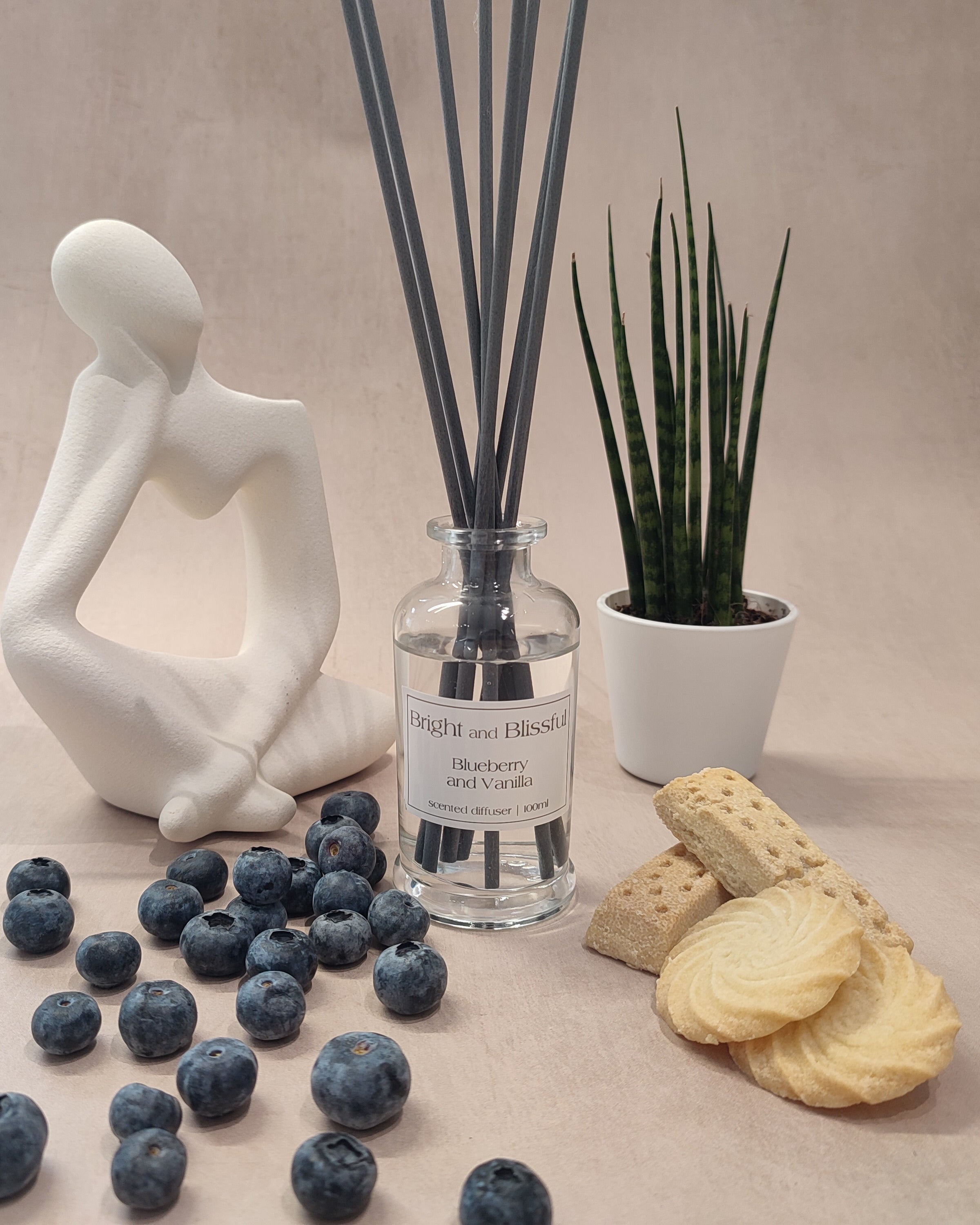 Reed diffuser with blueberries, cookies, and a decorative figure on a neutral background