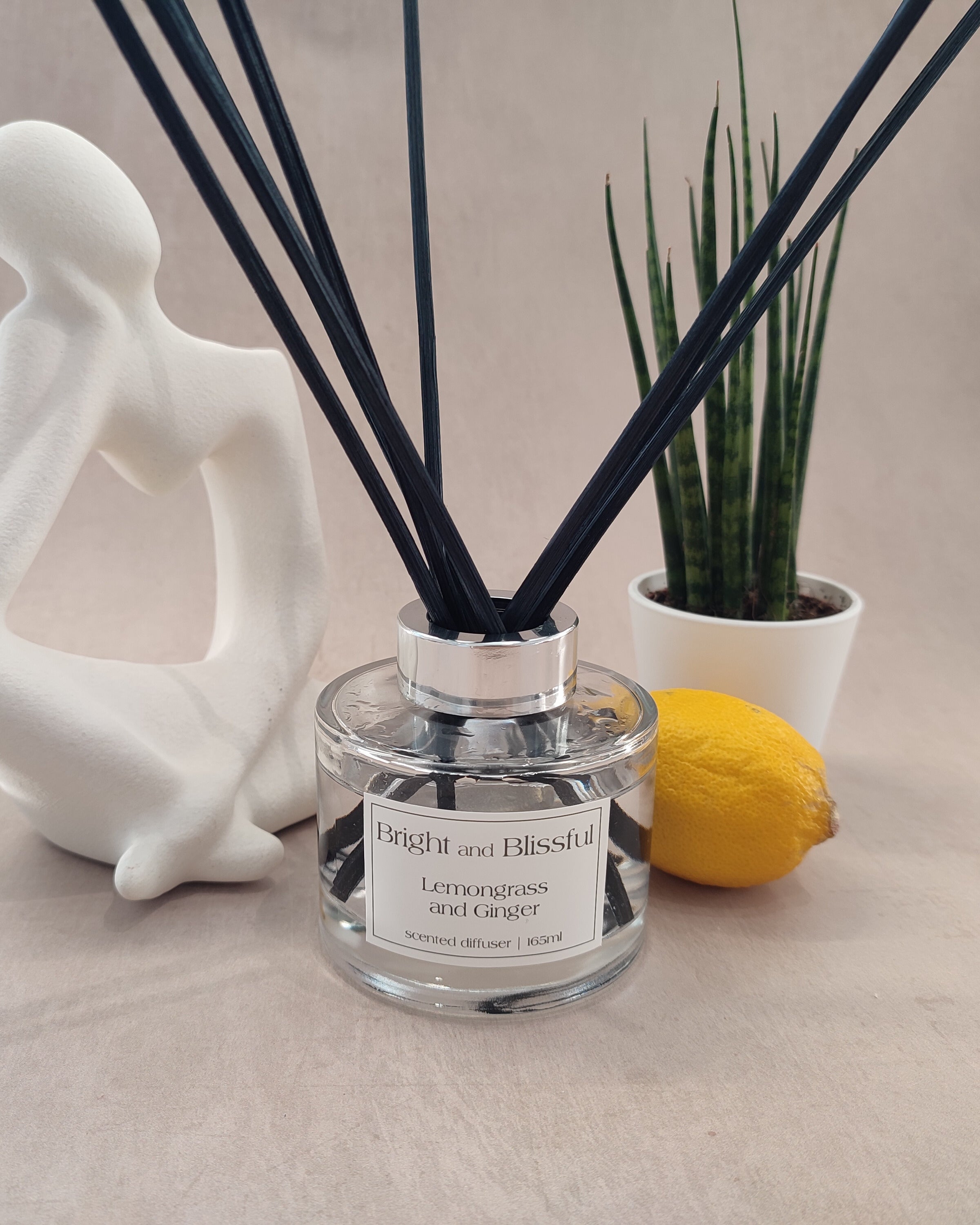 A diffuser with black reeds, a lemon, and a plant on a neutral background