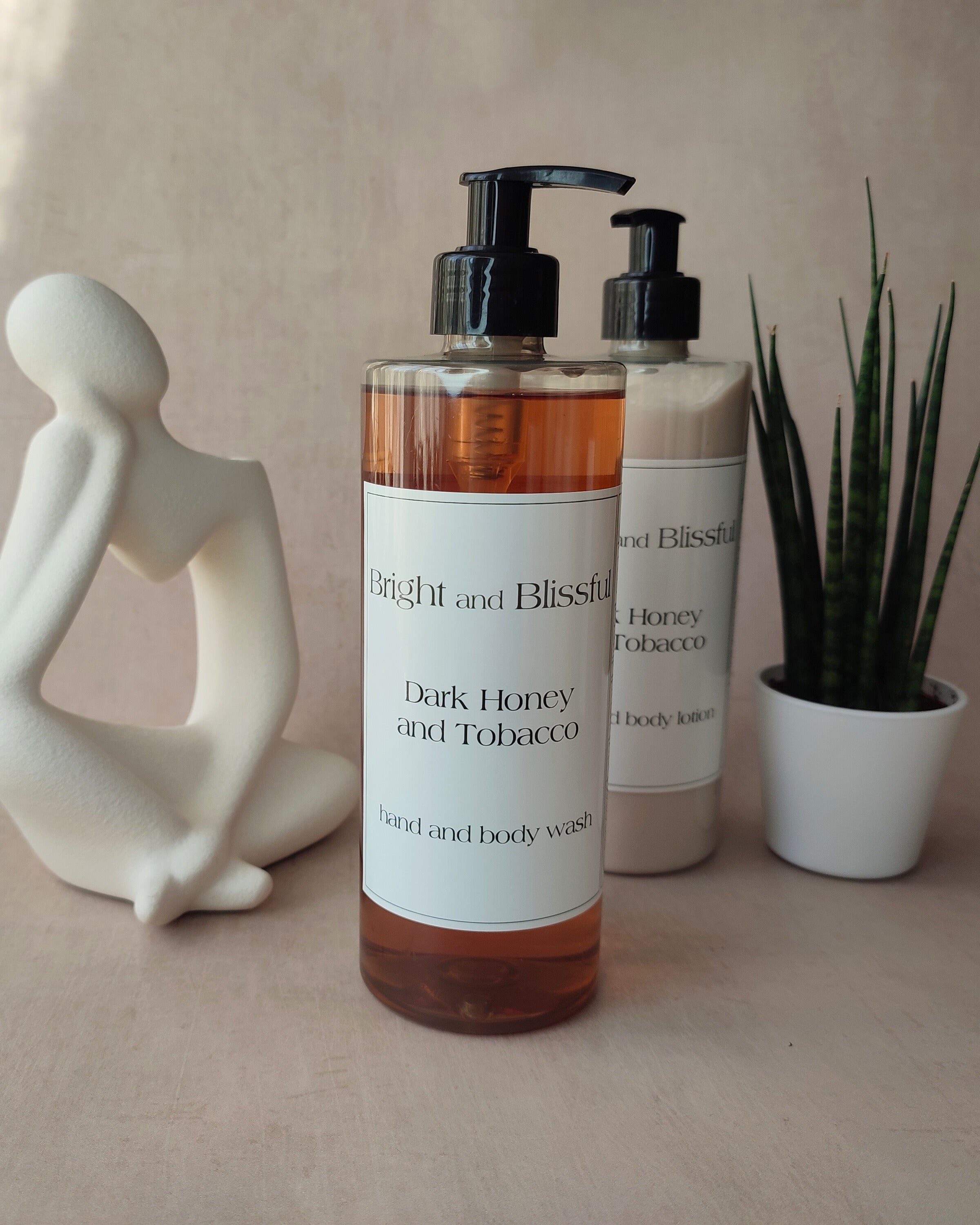 Two bottles of 'Bright and Blissful' body wash on a neutral background with a decorative figure and plant.