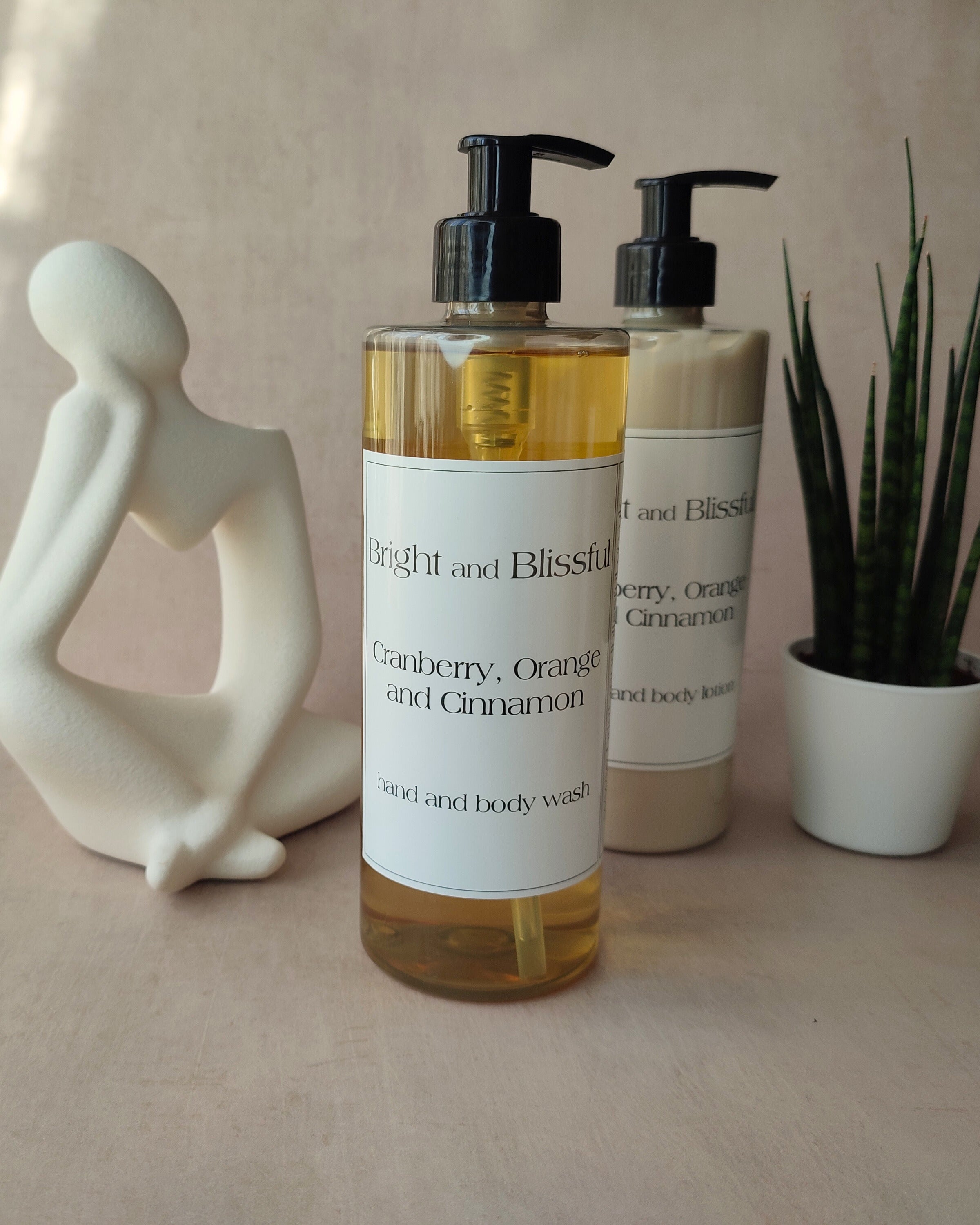 Two bottles of 'Bright and Blissful' body wash on a neutral surface with a plant and sculpture in the background.