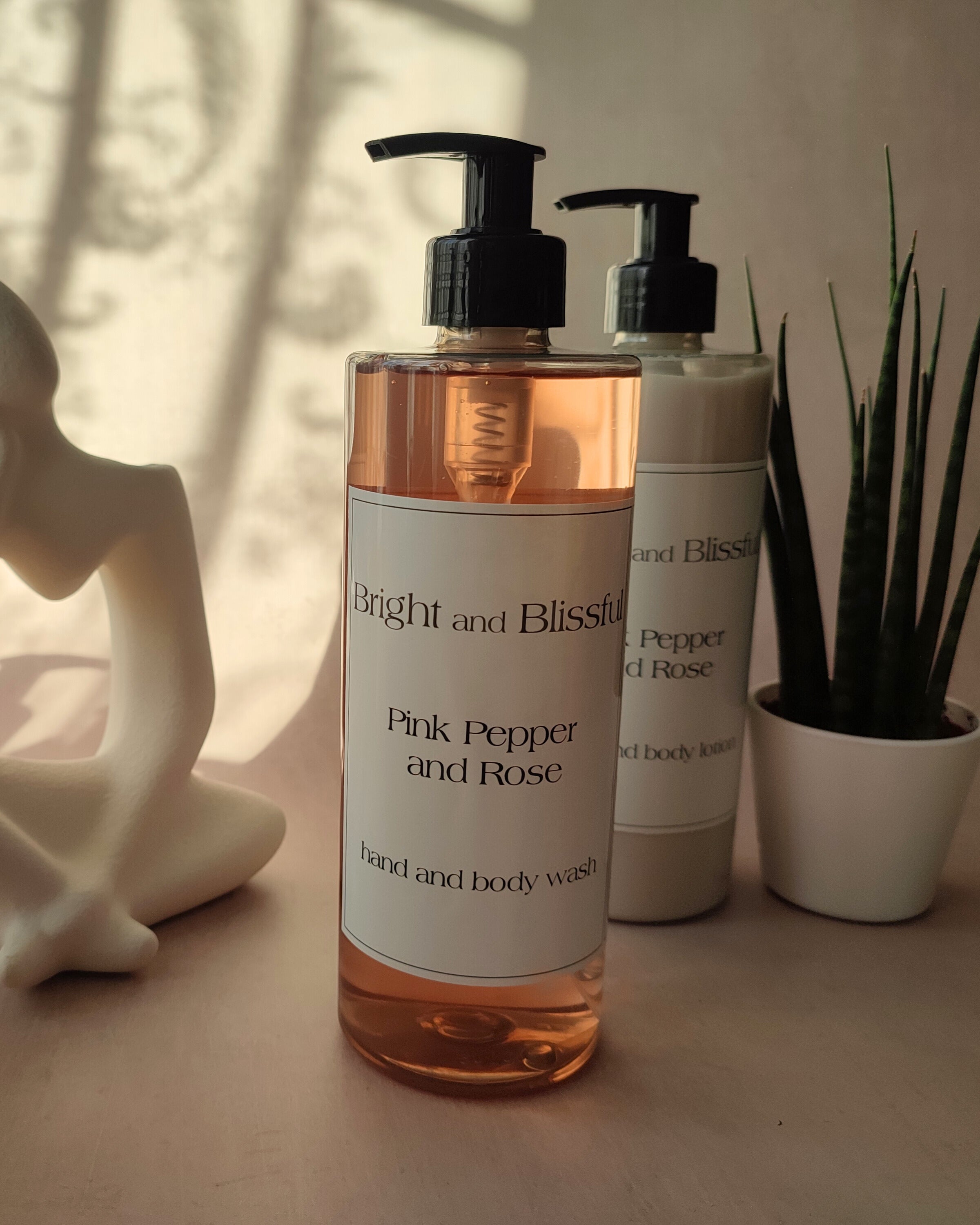 Bottle of 'Bright and Blissful' Pink Pepper and Rose hand and body wash on a surface with a plant and statue in the background.