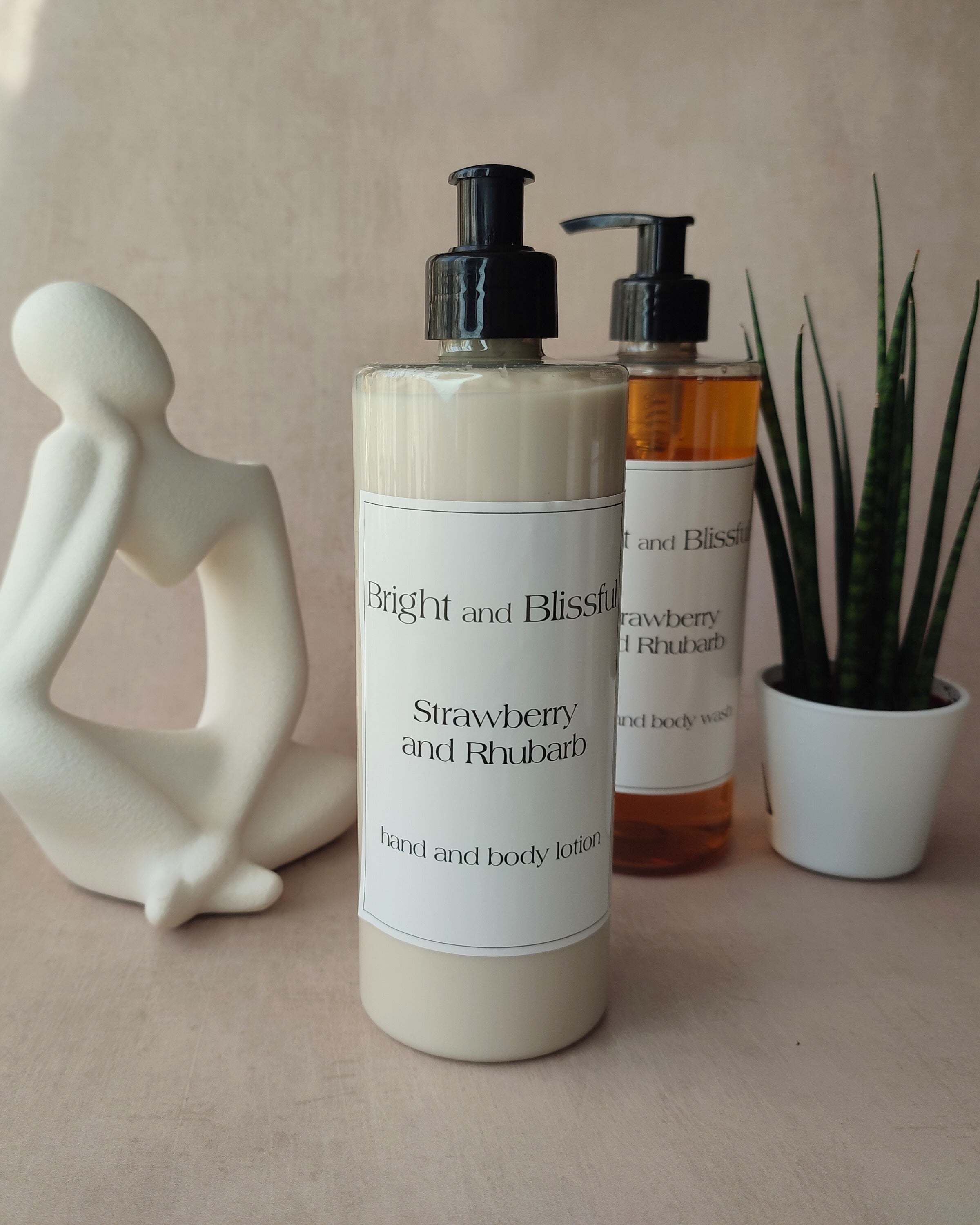 Two bottles of 'Bright and Blissful' Strawberry and Rhubarb hand and body lotion on a neutral background with a decorative figure and plant.