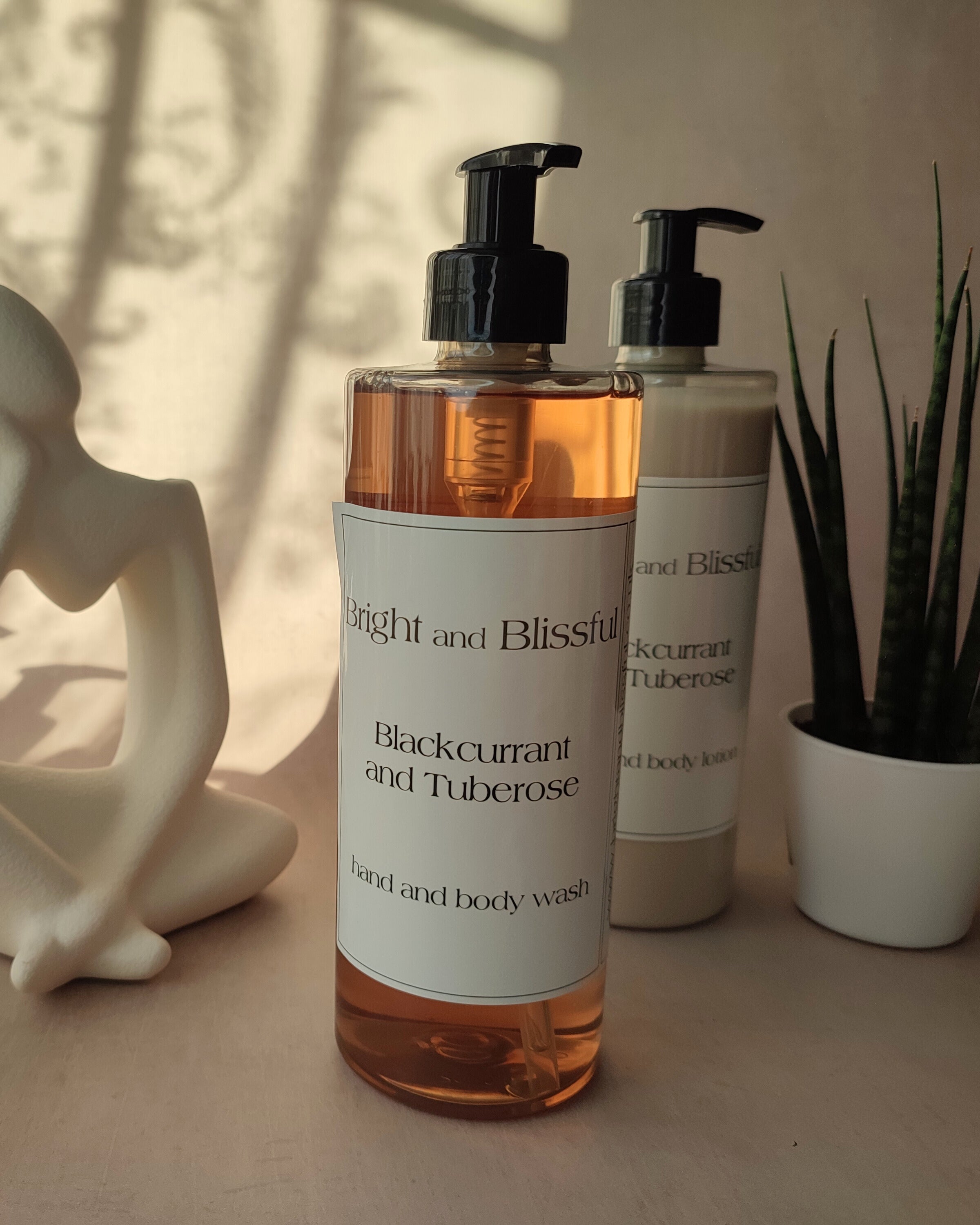 Two bottles of 'Bright and Blissful' hand and body wash on a surface with a plant and sculpture in the background.