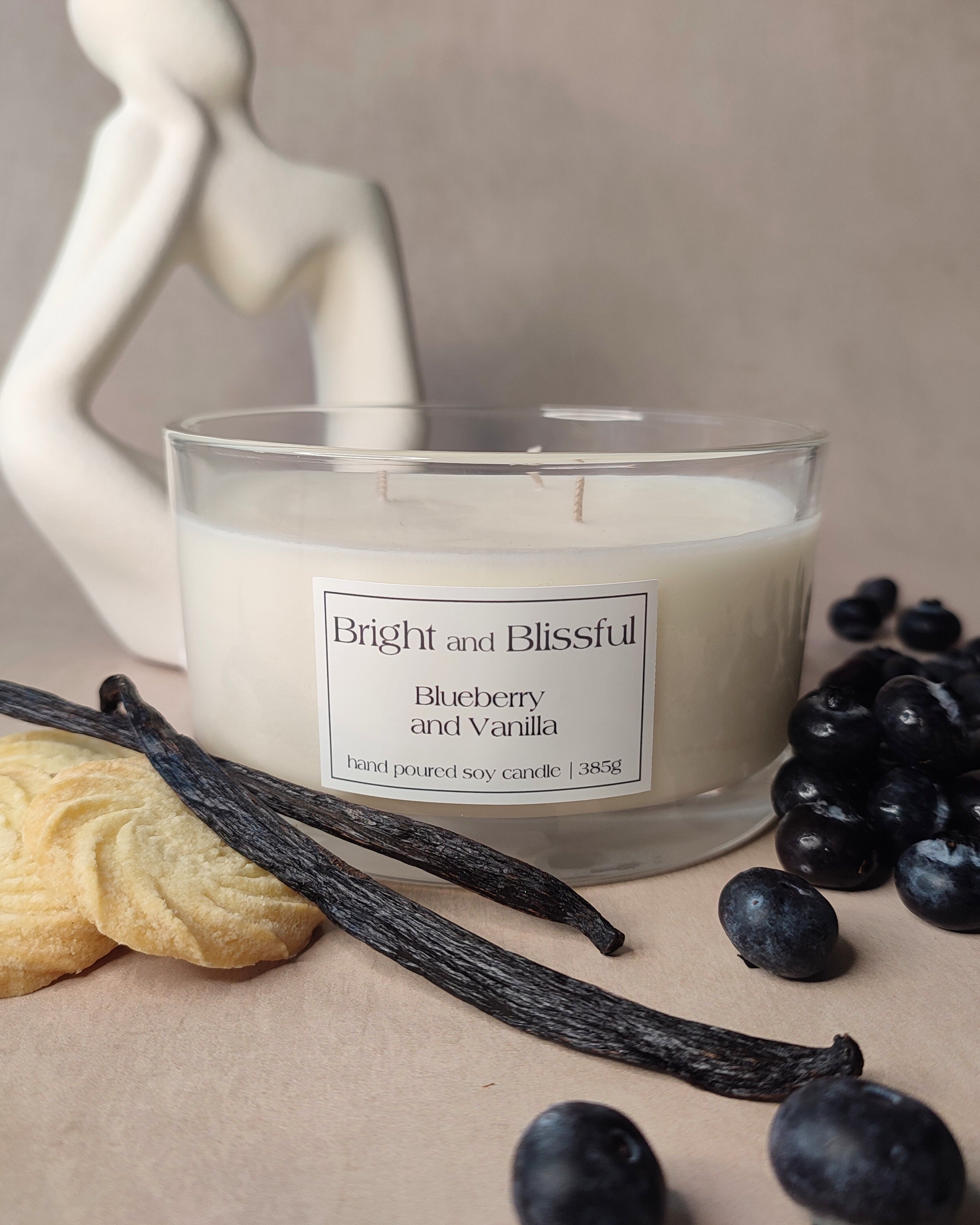 Candle labeled 'Bright and Blissful' with blueberries, vanilla beans, and a cookie on a neutral background