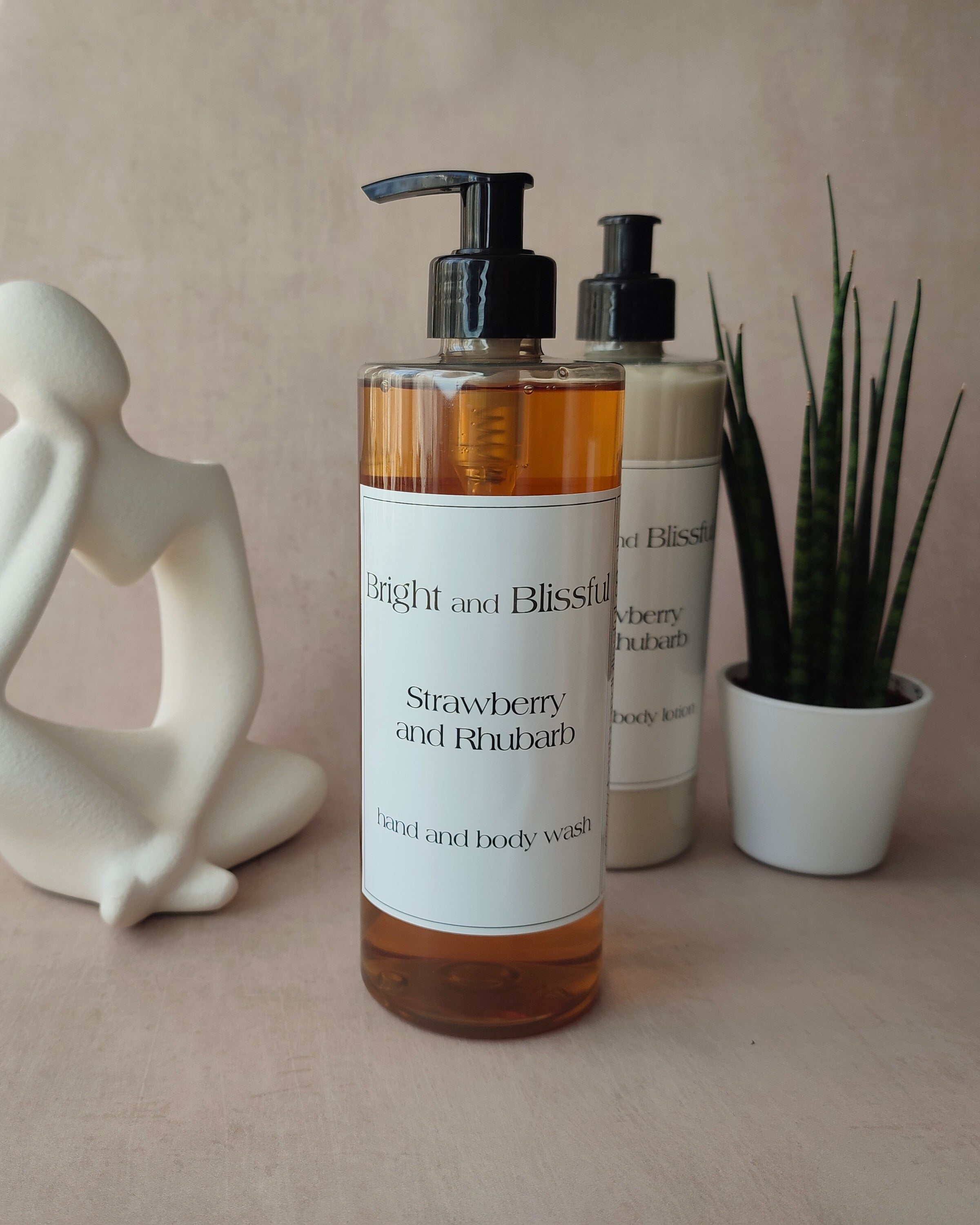 Two bottles of 'Bright and Blissful' strawberry and rhubarb body wash on a neutral background with decorative items.