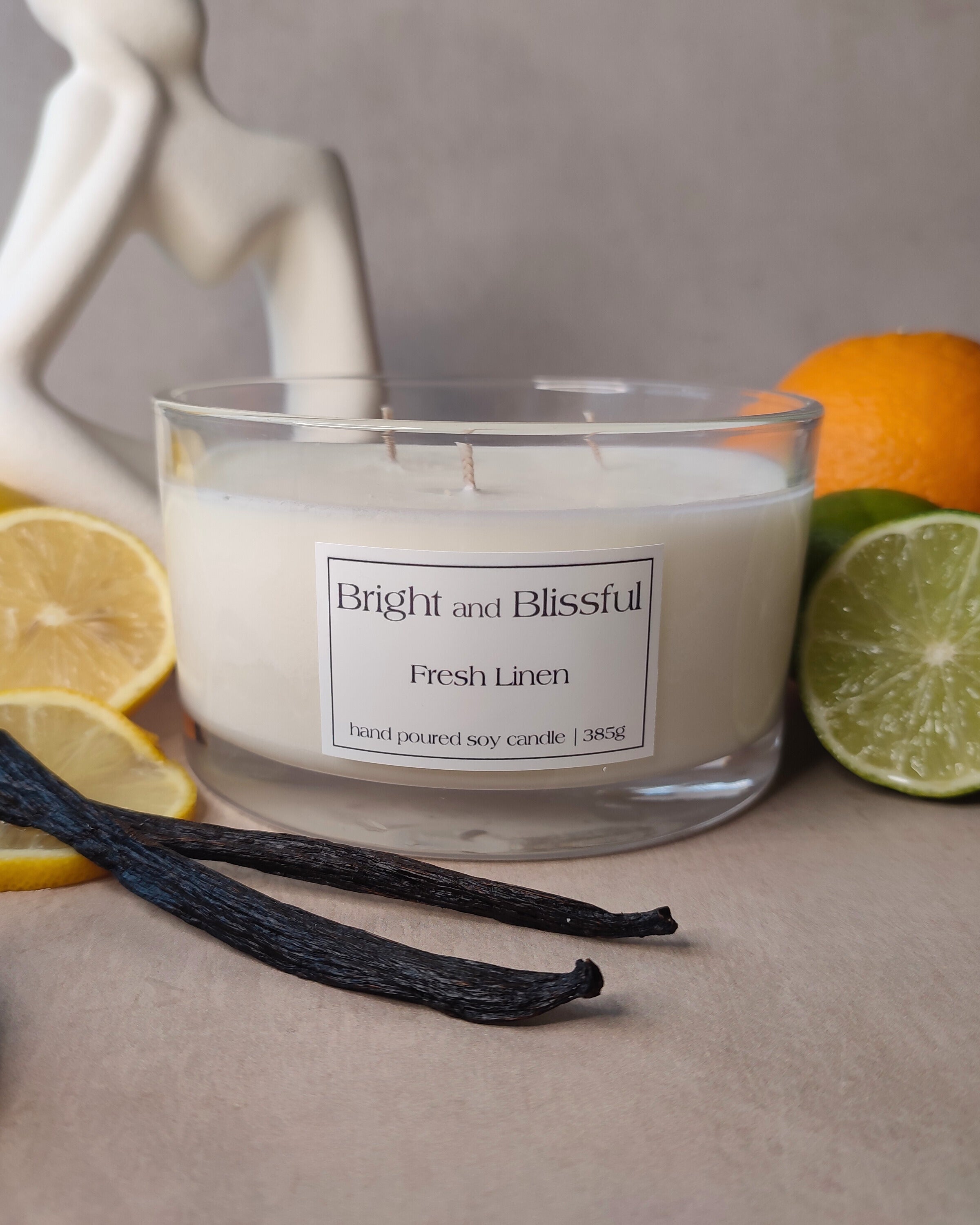 Candle labeled 'Bright and Blissful Fresh Linen' with lemons, oranges, and vanilla beans on a neutral background