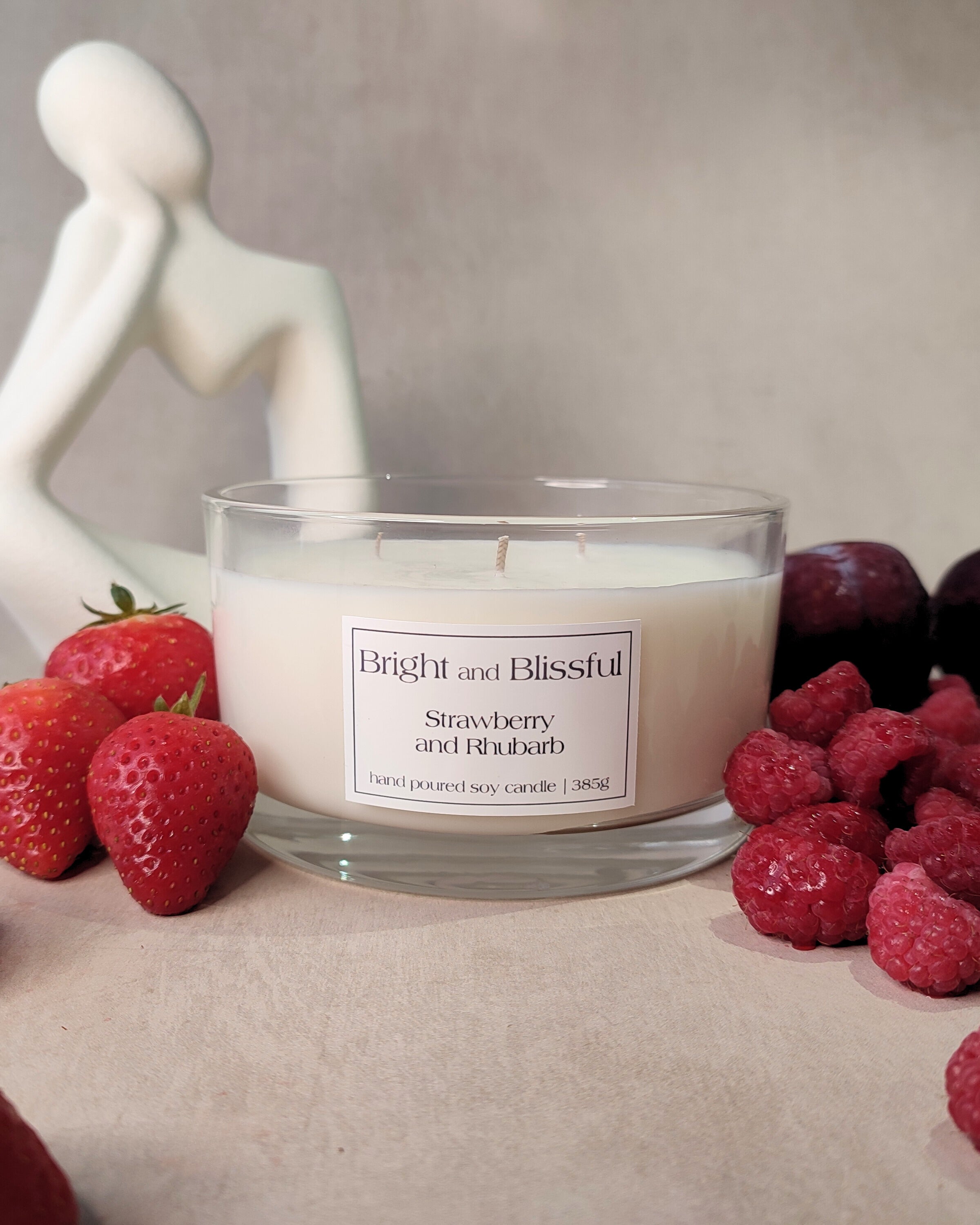 Candle labeled 'Bright and Blissful Strawberry and Rhubarb' surrounded by strawberries, raspberries, and a marble sculpture.