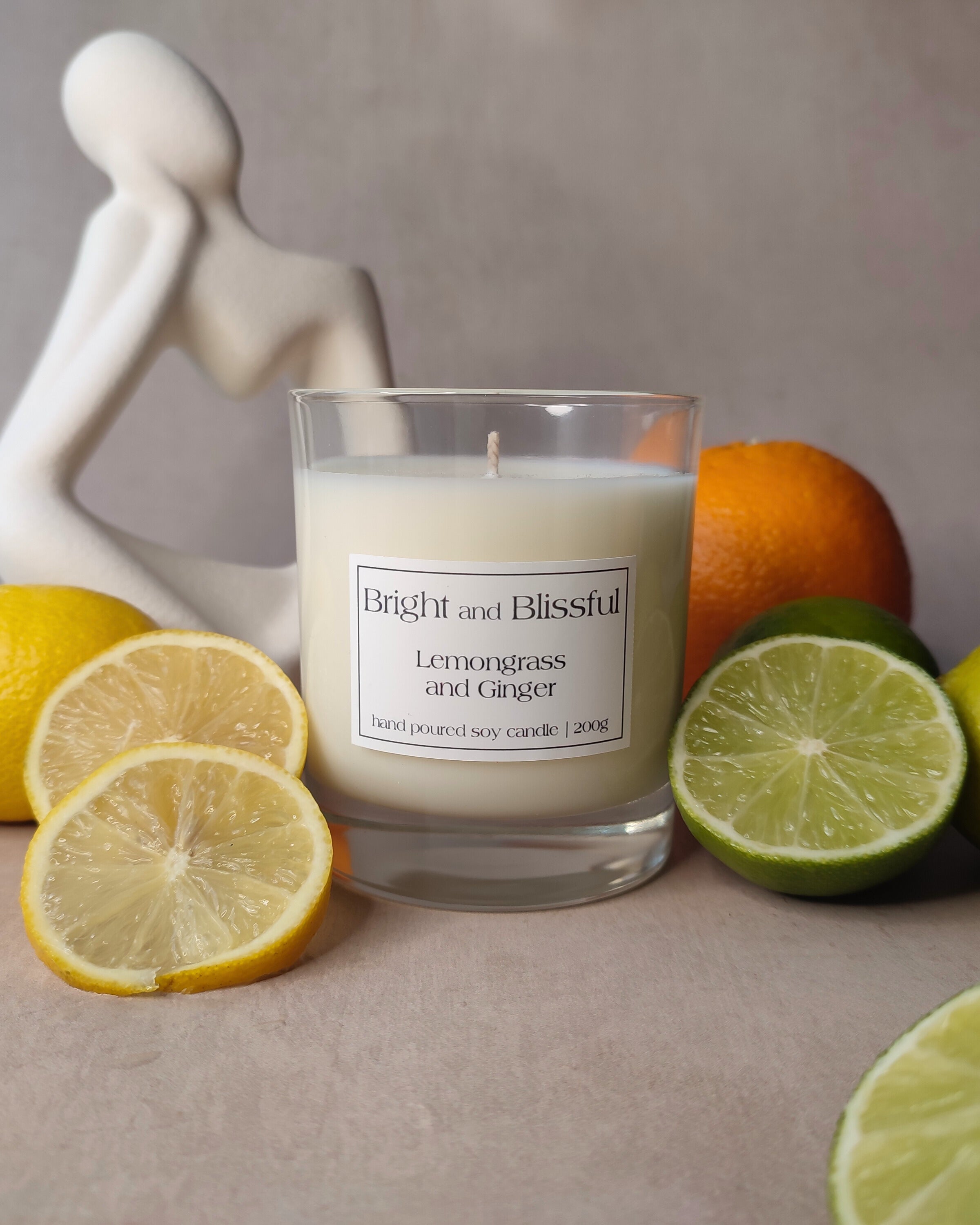 Candle labeled 'Bright and Blissful Lemongrass and Ginger' surrounded by lemons, limes, and an orange on a neutral background.