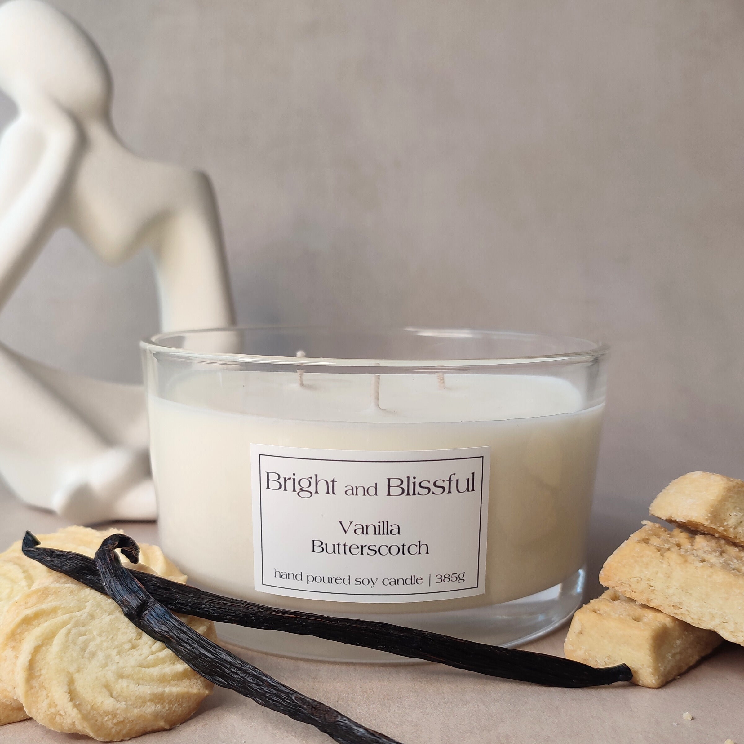 Candle labeled 'Bright and Blissful Vanilla Butterscotch' with cookies on a neutral background