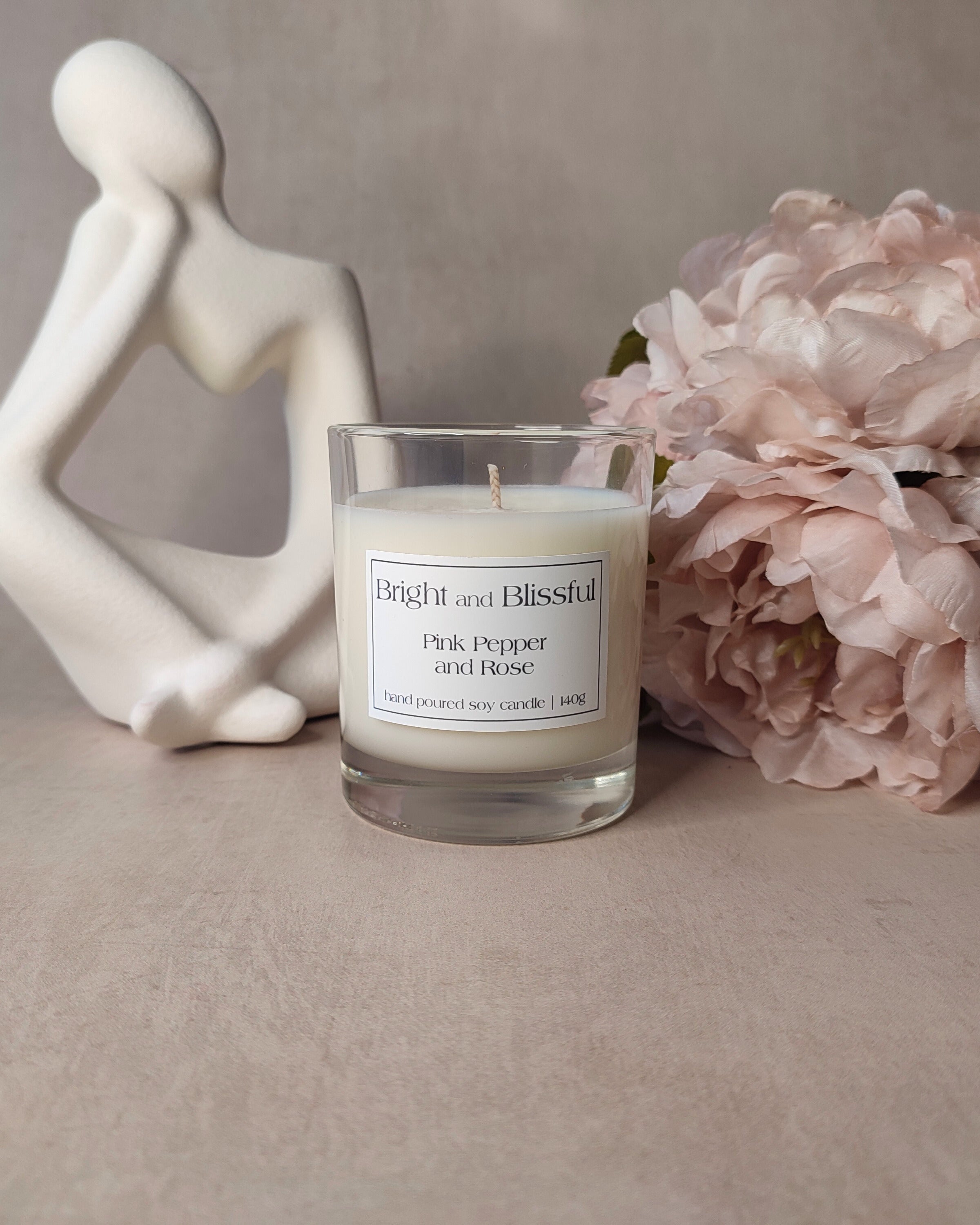 Candle labeled 'Bright and Blissful' with a sculpture and flowers on a neutral background
