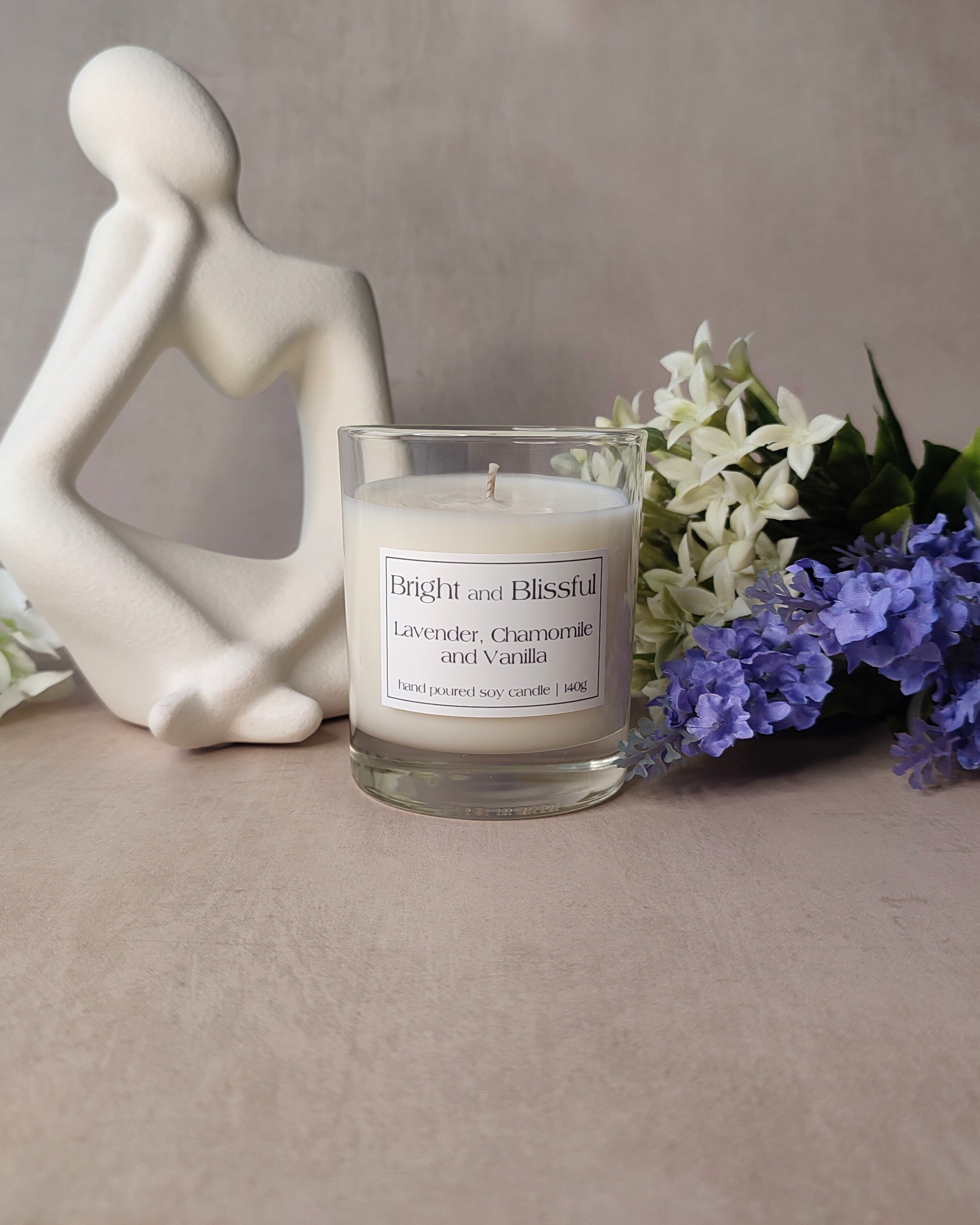 Candle labeled 'Bright and Blissful' with a decorative figurine and flowers on a neutral background