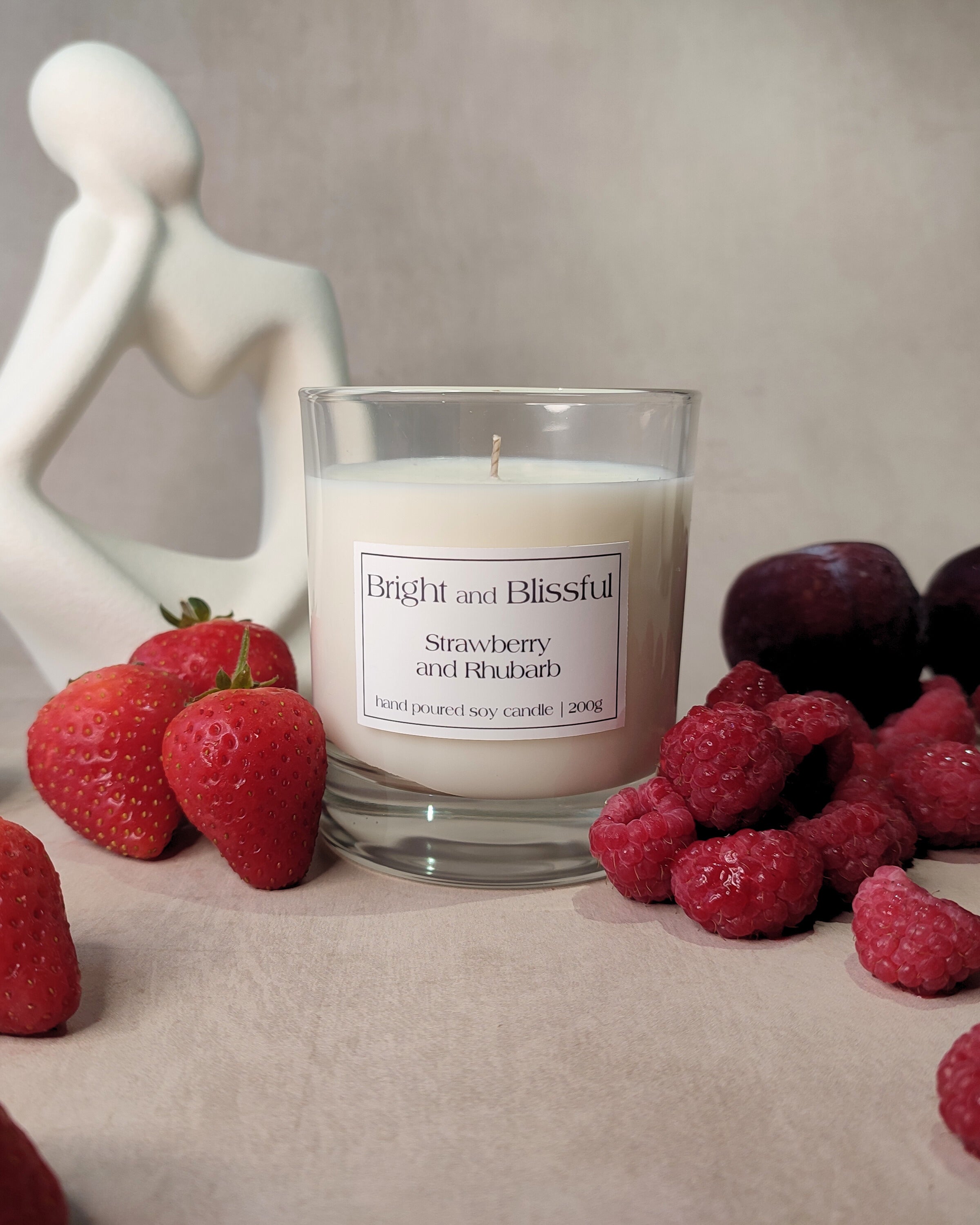 Candle labeled 'Bright and Blissful Strawberry and Rhubarb' surrounded by strawberries and raspberries on a neutral background.