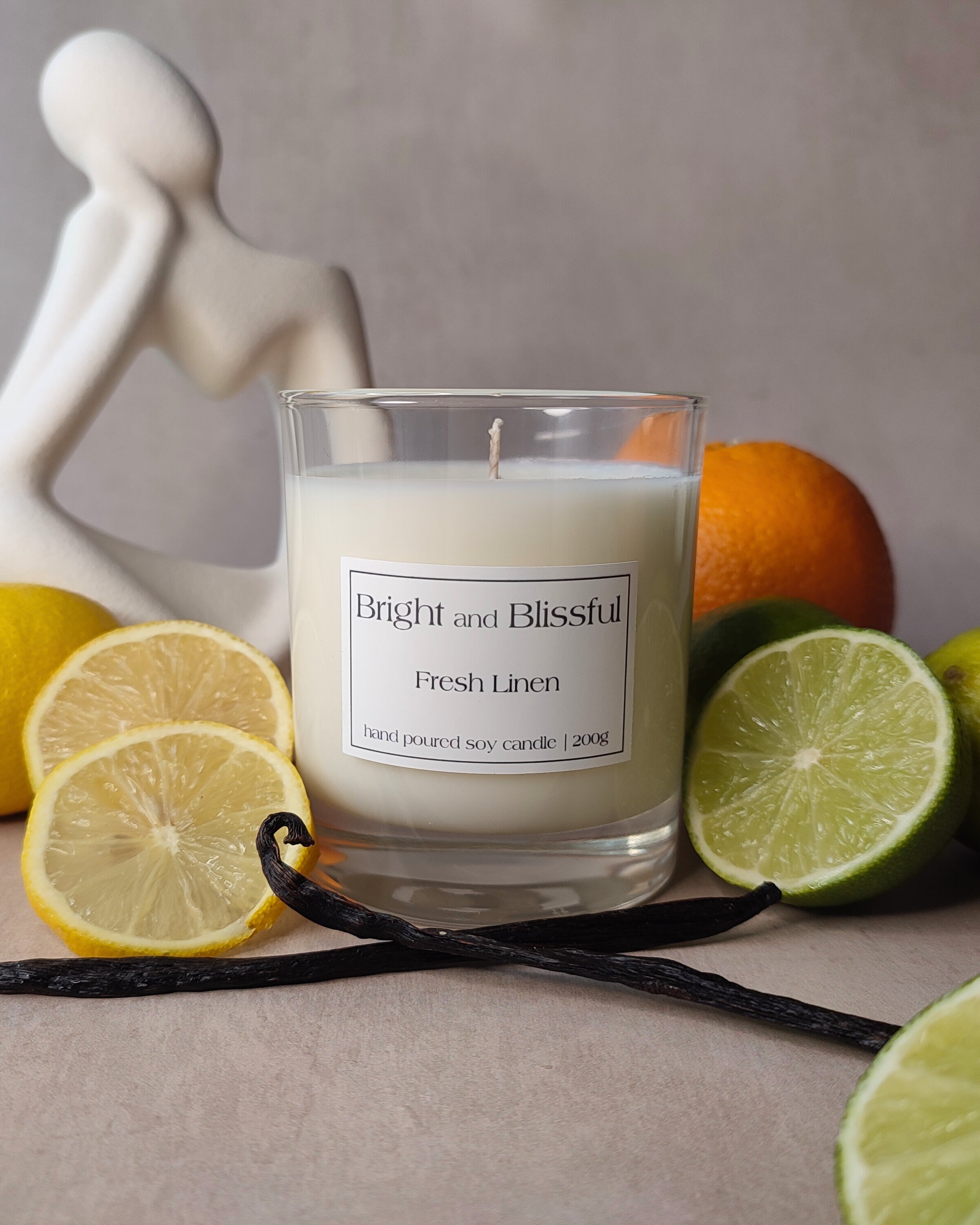 Candle labeled 'Bright and Blissful Fresh Linen' surrounded by lemons, limes, and an orange on a neutral background.