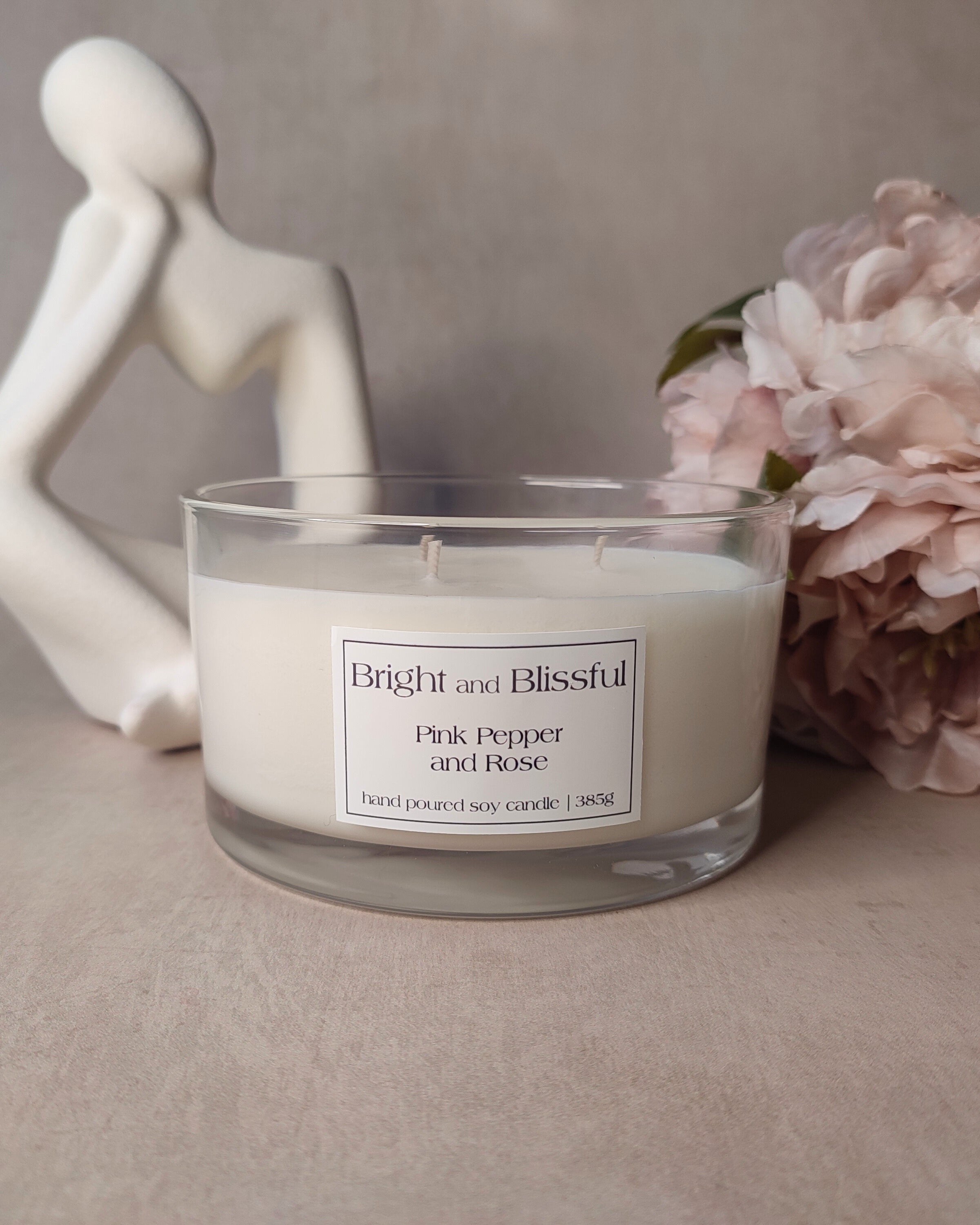 Candle in a glass jar with a label, next to a decorative figurine and flowers on a neutral background