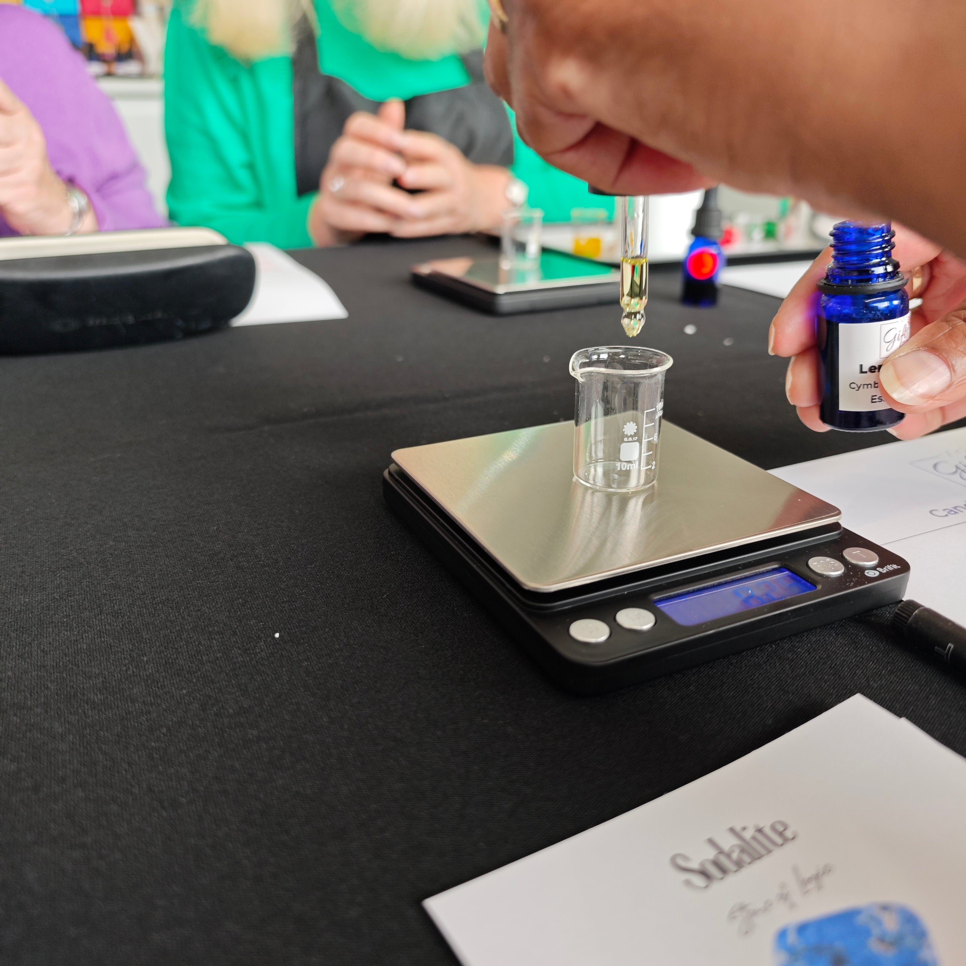 Person using a dropper to add liquid to a small container on a digital scale with another person in the background.