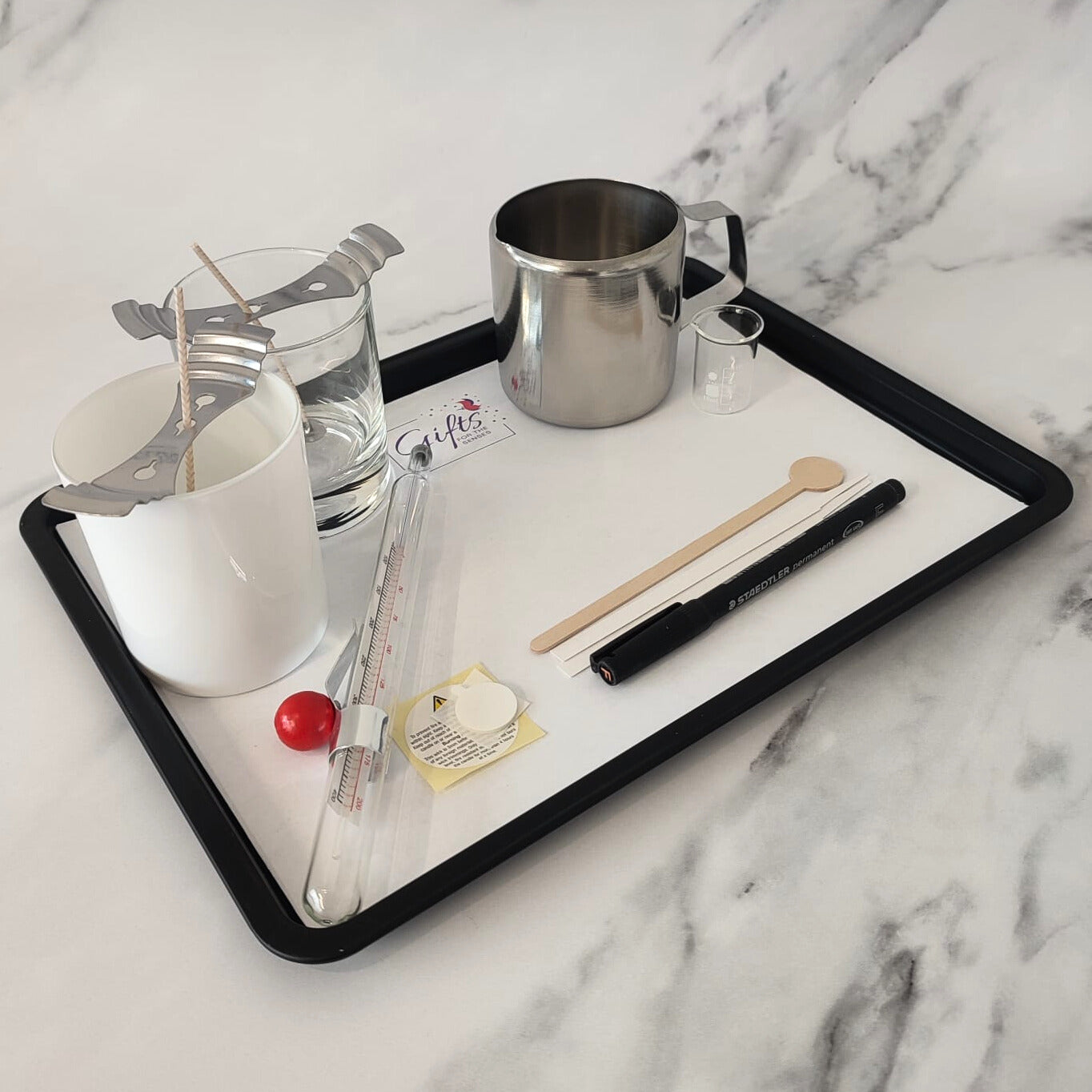 Tray with candlemaking tools including a water glass, thermometer, and various utensils on a marble surface.