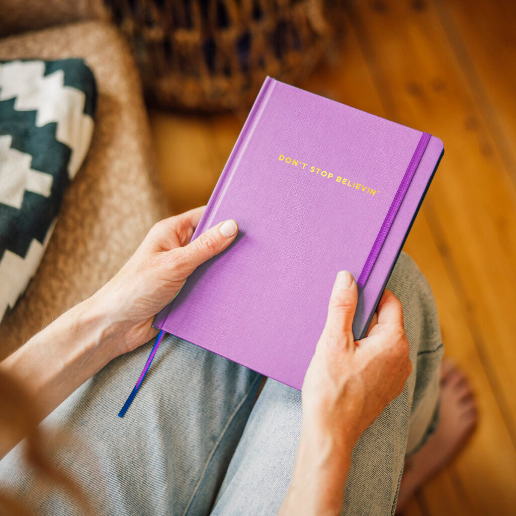 Purple notebook titled 'Don't stop believin' held by a person on a wooden floor.