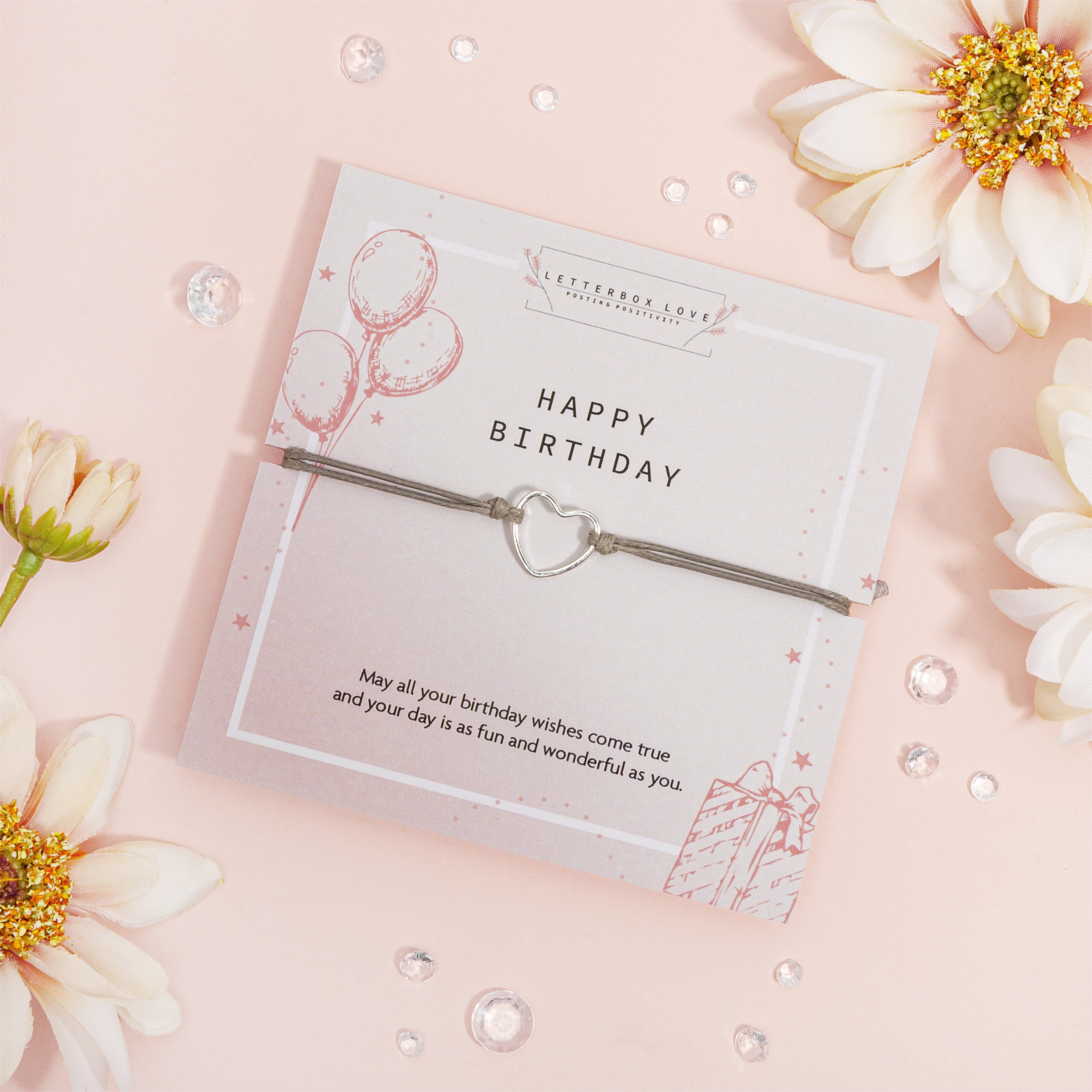 Silver bracelet with heart charm in a 'Happy Birthday' card packaging on a pink background with flowers.
