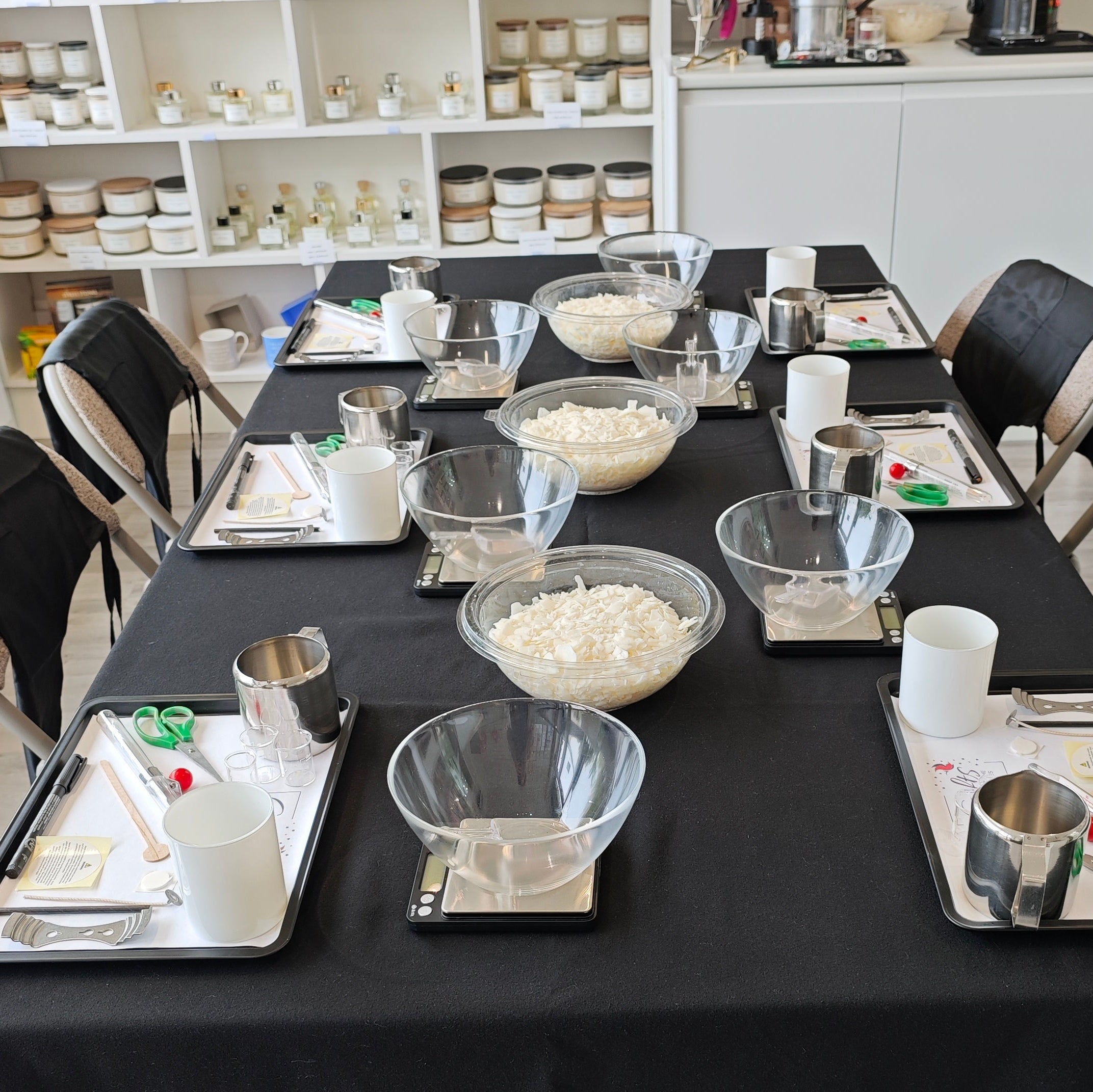 Table set for a class with various items on a black tablecloth in a room with shelves and a double boiler for candle making.