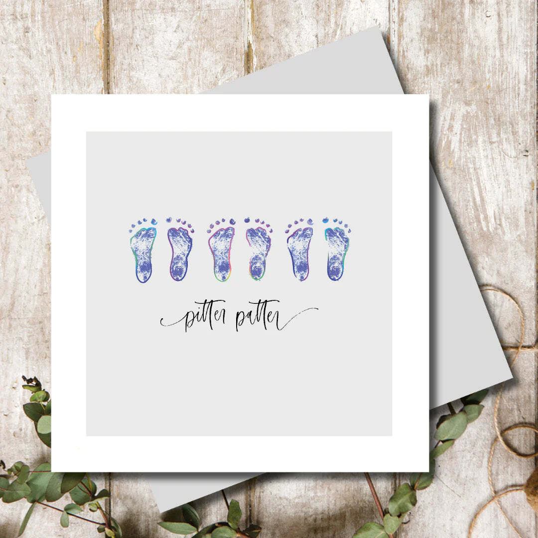 Greeting card with watercolor footprints and 'pitter patter' text on a wooden surface.