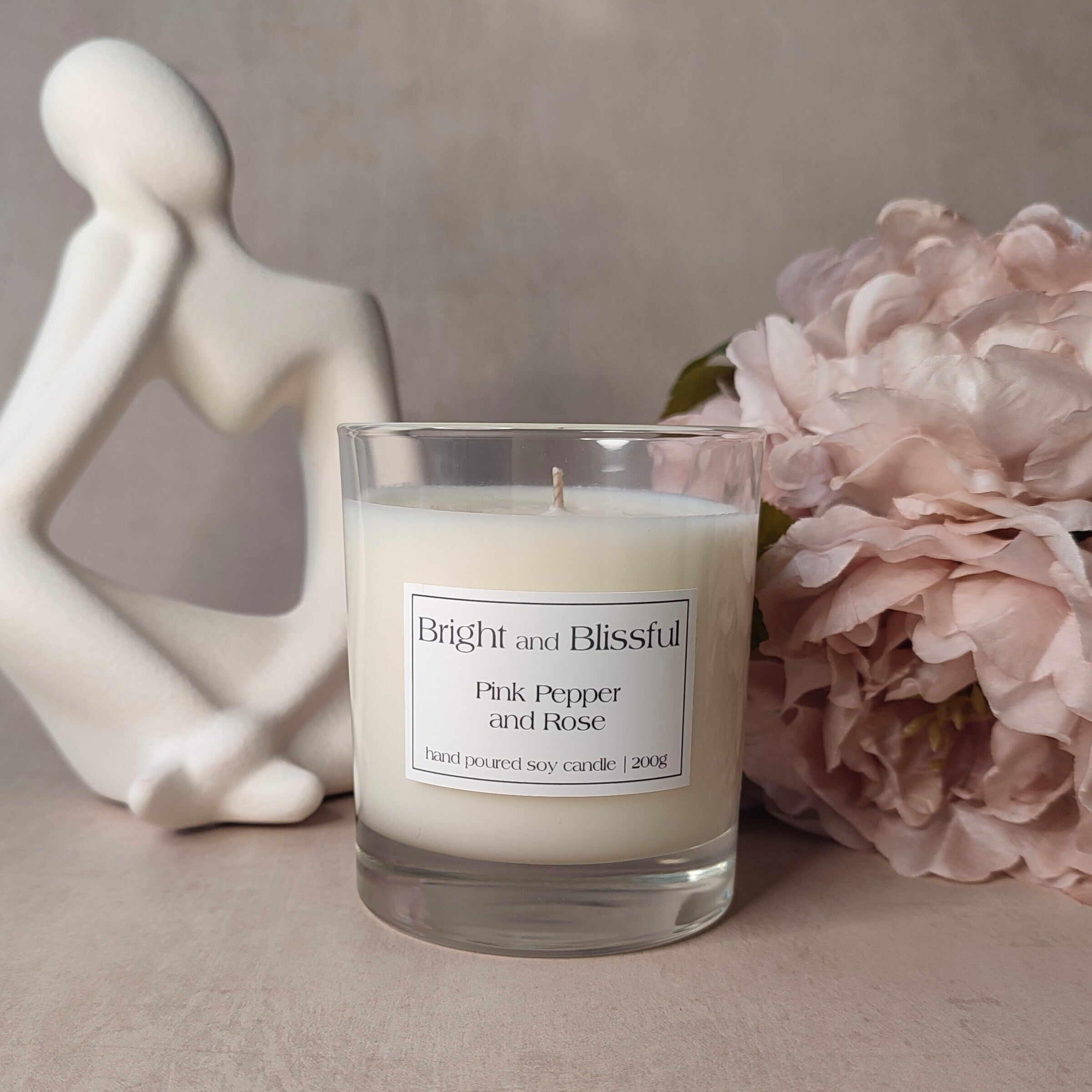 Candle labeled 'Bright and Blissful Pink Pepper and Rose' with a decorative figurine and flowers on a neutral background