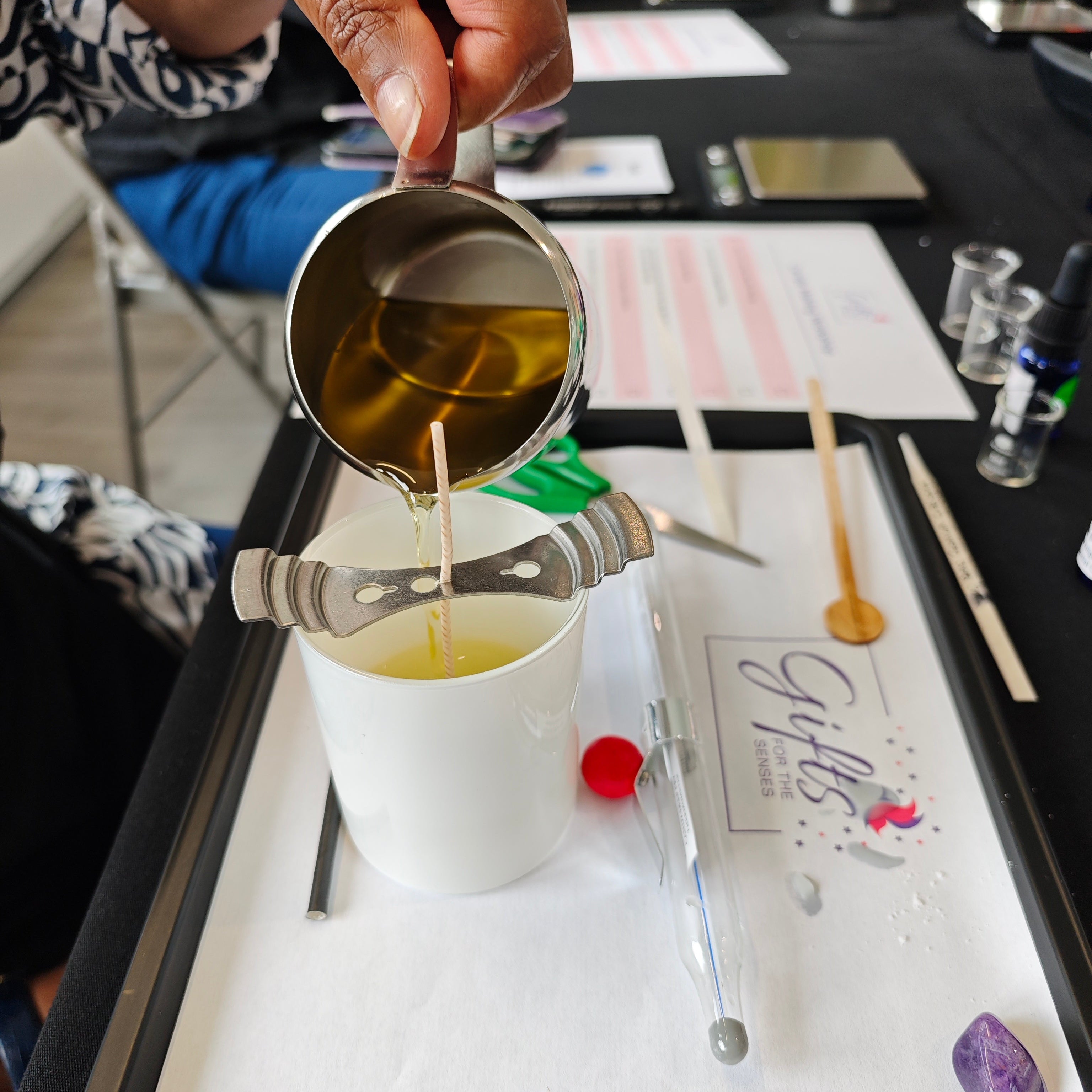 Hand pouring liquid from a small metal jug into a white container on a tray with various items.