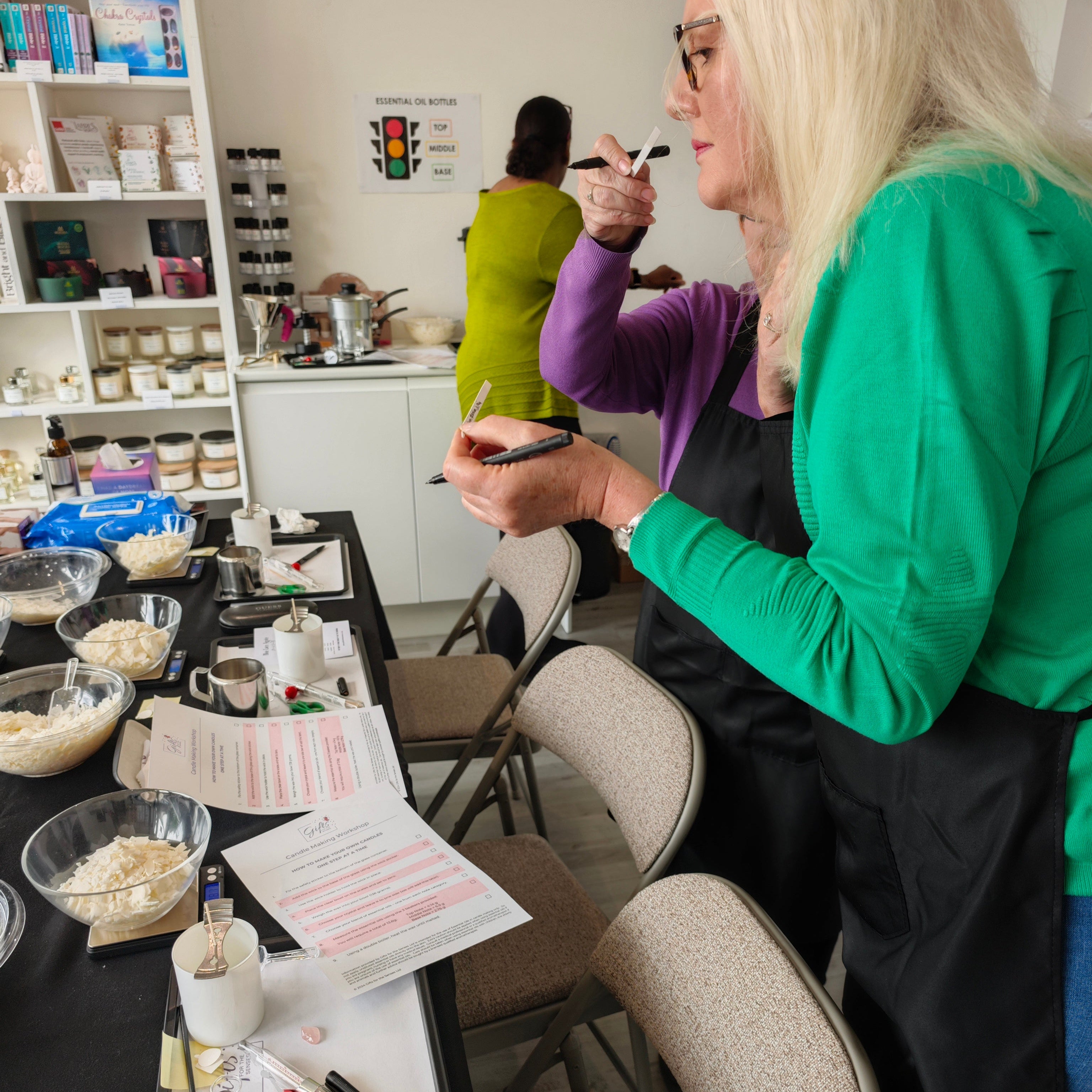 Woman in a green shirt and black apron at a candle making class sniffing essential oils.