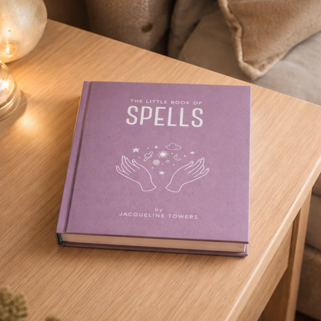 Purple book titled 'The Little Book of Spells' by Jacqueline Towers on a wooden table.