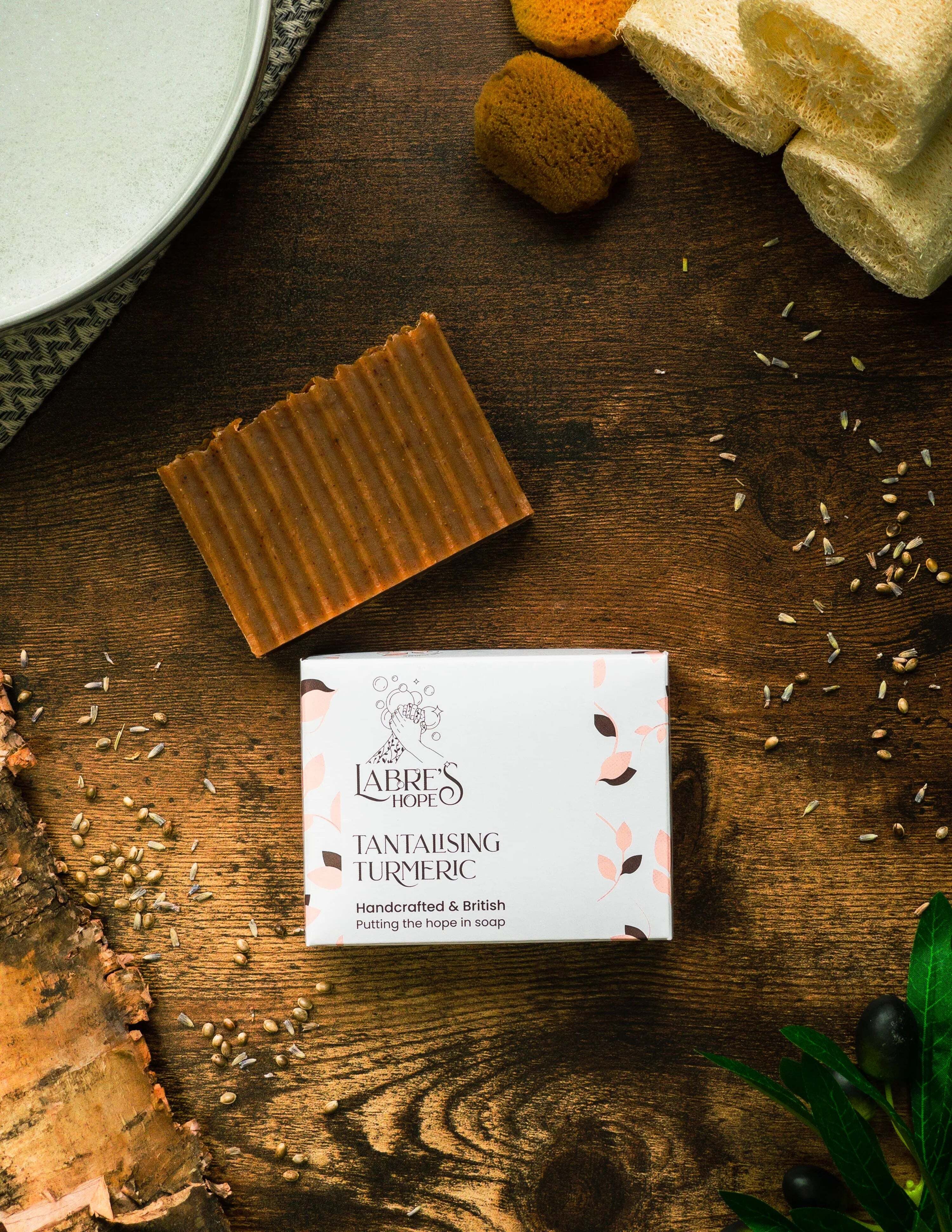 Bar of soap and its packaging on a wooden surface with herbs and a towel.