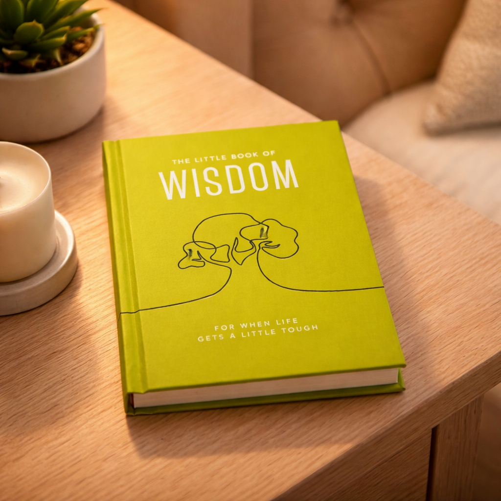 Green book titled 'The Little Book of Wisdom' on a wooden table with a cup and plant in the background.