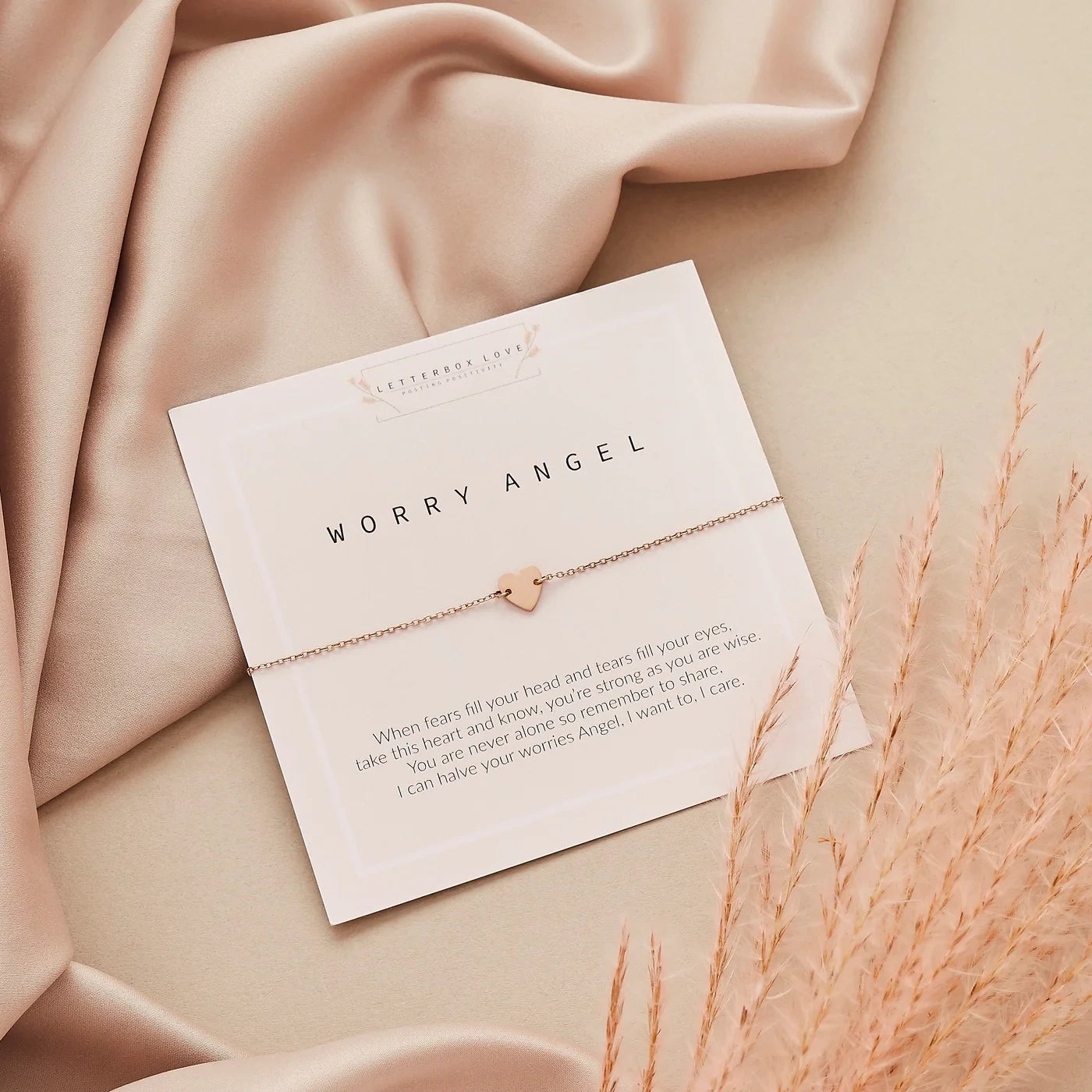 Rose gold 'Worry Angel' necklace on a card with text, placed on a soft pink fabric background.